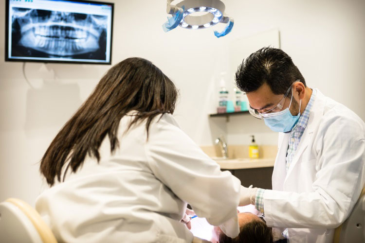 A dental professional in a lab coat and stethoscope, performing a procedure on a patient s teeth while another person observes.