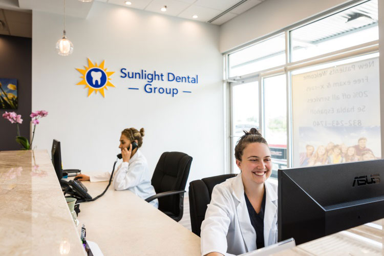 A dentist s office interior with a woman at the reception desk and two individuals working in the background.