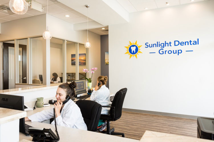 The image shows an interior view of a dental office, featuring a reception area with a large sign that reads  SUNLIGHT DENTAL GROUP.  There are two individuals present  one appears to be seated at the reception desk and the other is standing behind it. The environment includes a waiting area with seating, a receptionist s counter, and a wall-mounted sign indicating the dental practice s name.