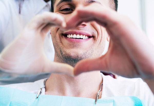 A man in a dental chair, holding up a heart sign with his hands, smiling broadly at the camera.