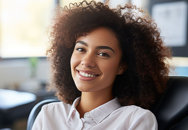 The image is a photograph of a smiling woman with curly hair, wearing a white blouse and sitting in an office environment.