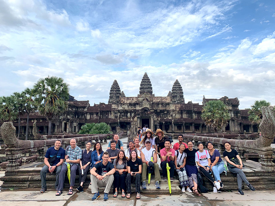A group of people posing for a photo in front of the Angkor Wat temple complex, with palm trees and a clear sky visible.