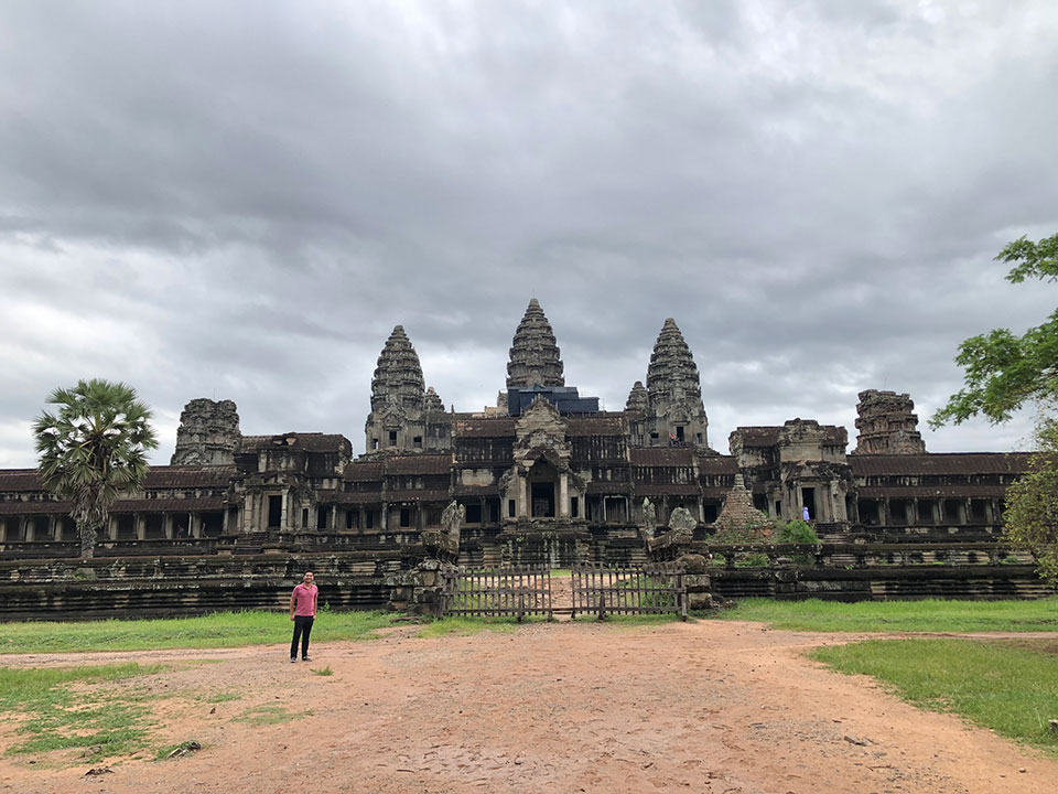 The image features the grand entrance to the Angkor Wat temple complex in Cambodia, with a person standing in front of the impressive stone structures under an overcast sky.