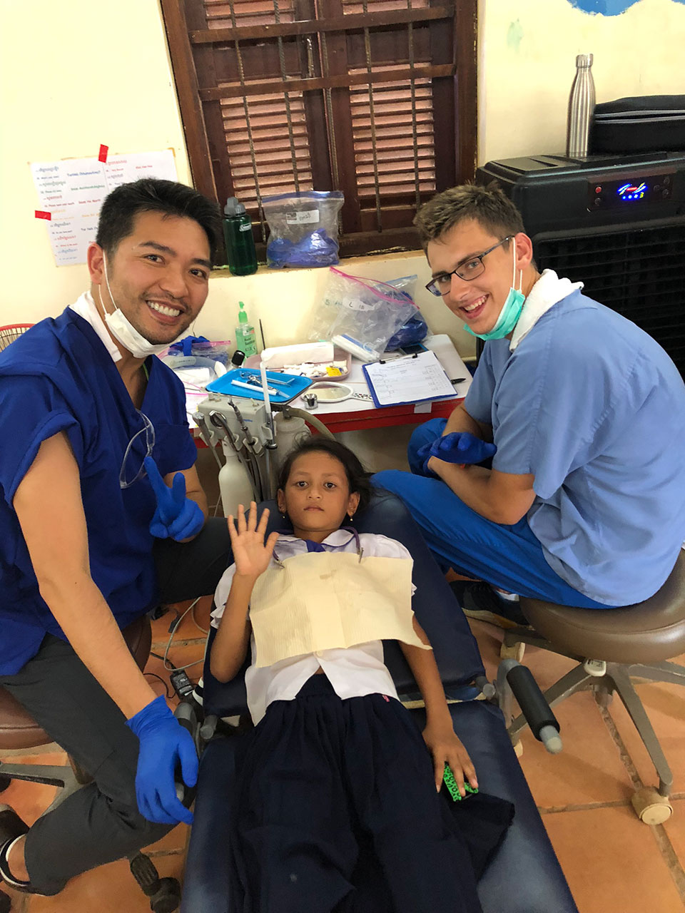 In the image, three individuals are present in a medical setting. A young child is lying on a bed with medical equipment nearby, and two adults are standing next to the bed, one of whom appears to be a healthcare professional given their attire and the presence of medical supplies. The environment suggests a clinical or hospital-like atmosphere.