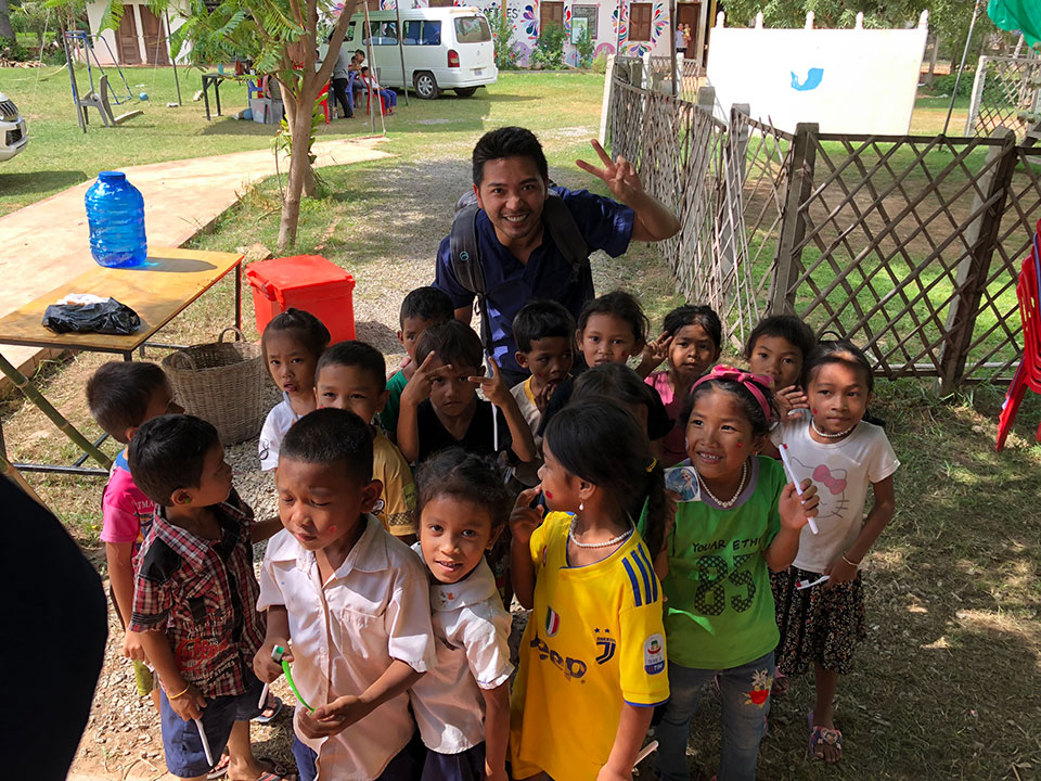 The image shows a group of children standing in front of an adult, with the adult waving at the camera. They are outdoors, possibly in a schoolyard or park, as suggested by the presence of trees and a fence in the background.
