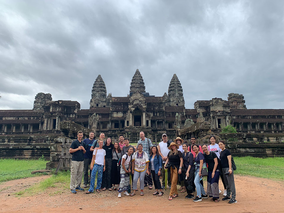 The image is a photograph of a group of people posing in front of an ancient temple complex, Angkor Wat, with the sky and clouds in the background.