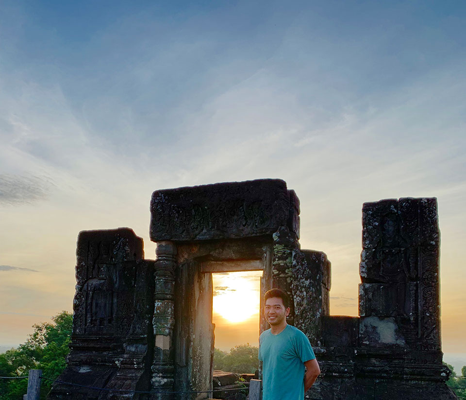 The image features a person standing in front of an ancient, stone temple entrance with a sunset or sunrise in the background.