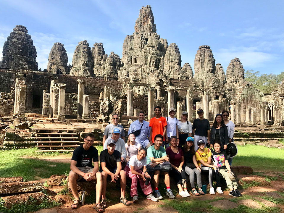 A group of people posing in front of ancient stone ruins, with a large temple-like structure featuring multiple towers and intricate carvings.