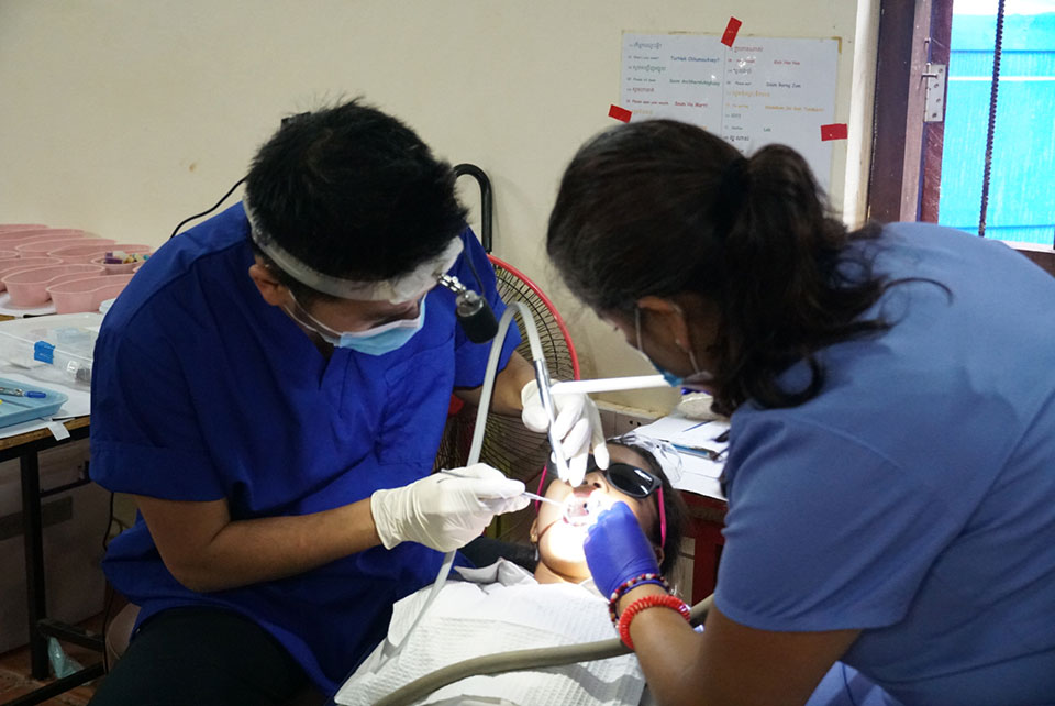 Two healthcare professionals performing a medical procedure, with one individual wearing surgical gloves and the other using a dental drill on a patient's tooth.
