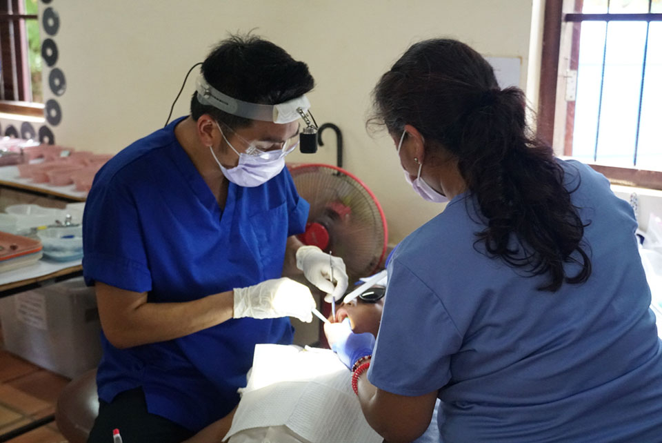 A medical professional in a blue uniform is assisting another individual with a medical procedure, both are wearing gloves and the scene appears to be set in an indoor healthcare facility.