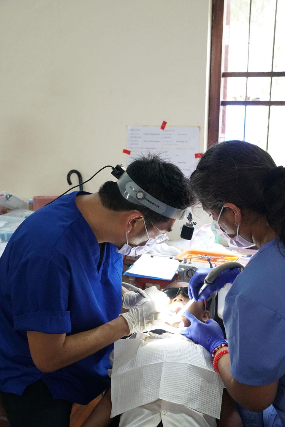 The image shows a medical professional performing dental work on a patient, with the dentist using a drill and wearing protective eyewear.