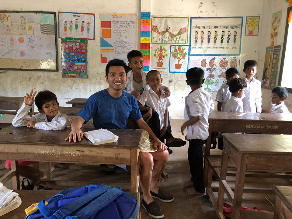 In the image, a man is standing in front of a classroom full of students. He appears to be engaged in an activity or conversation with them.