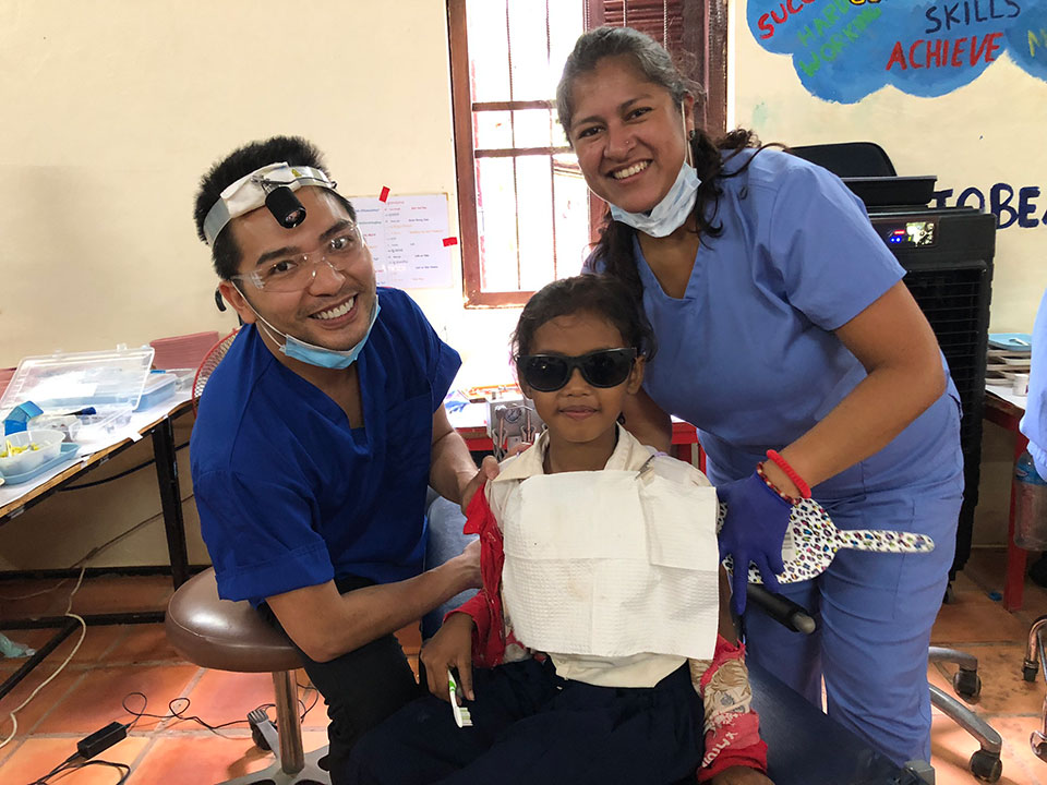 The image shows a group of people, including two individuals wearing medical masks and goggles, standing around a young child in a wheelchair. They are posing for the camera with smiles on their faces.