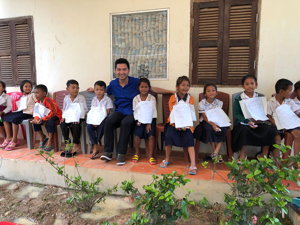 The image shows a group of children sitting in front of an adult, who appears to be supervising them. They are all seated on a bench, and the setting seems to be outdoors with a building in the background.