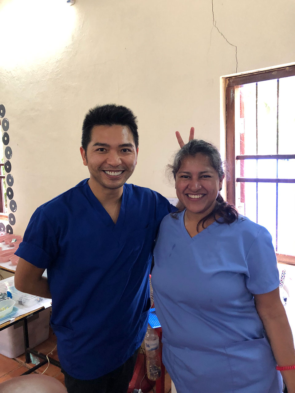 Two individuals, one male and one female, both dressed in professional attire, standing together with a smile, in what appears to be a clinical or educational setting.