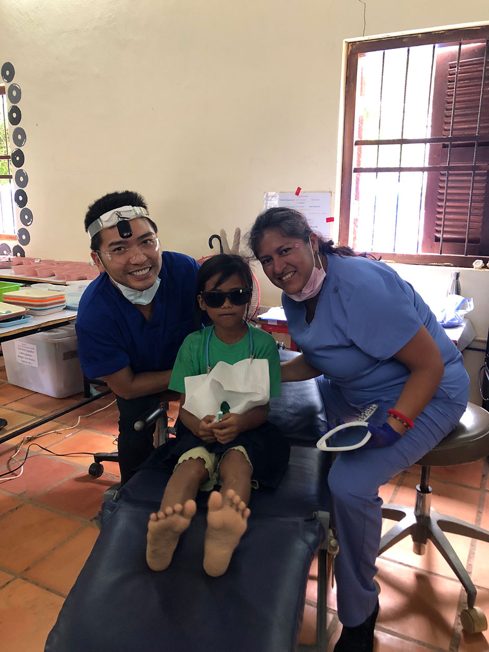 The image shows a medical setting with three individuals: a young child lying on a bed, an adult seated at the foot of the bed, and another adult standing by a desk. The room appears to be a hospital or clinic, indicated by the presence of medical equipment and personnel.