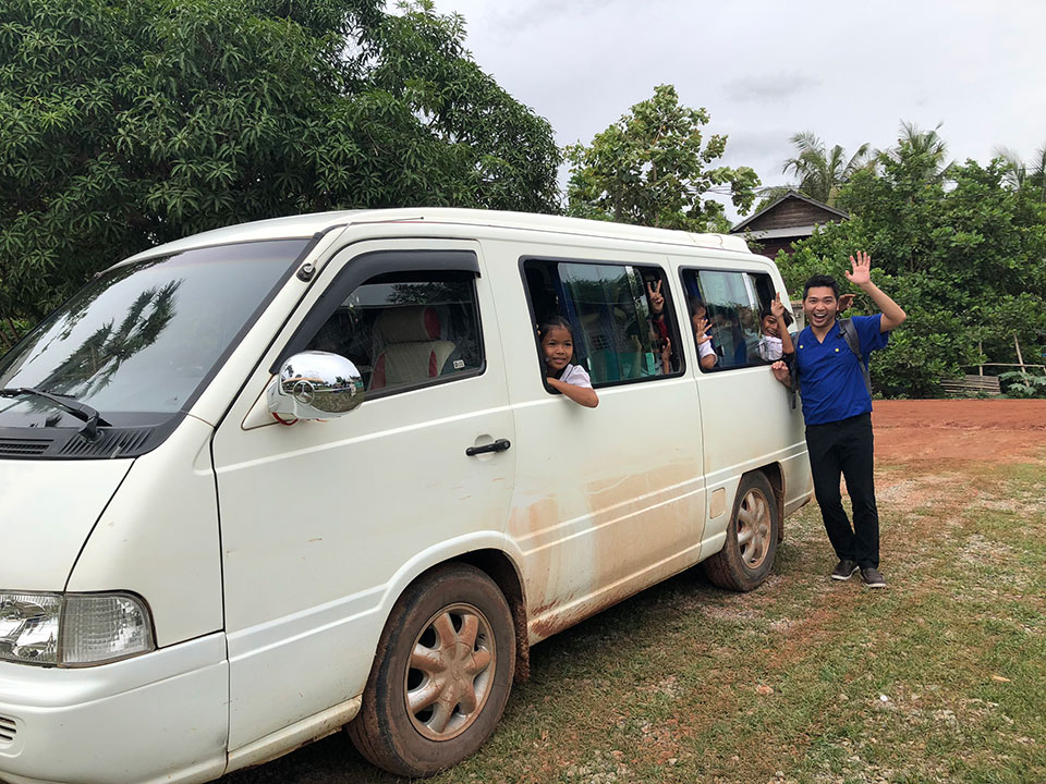 The image depicts a white van with tinted windows, parked on a dirt road. People are standing in front of the vehicle, waving and smiling at the camera.