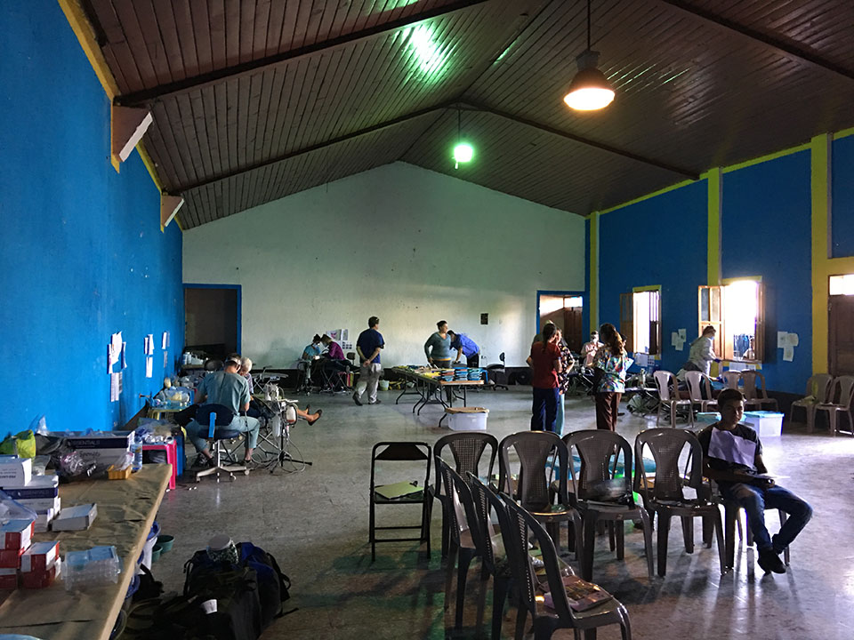 The image shows an interior space, likely a church or hall, with rows of chairs facing a central area where several people are standing and interacting. There is a large open space in the background with a blue wall and a ceiling fan. Various items, possibly medical supplies or equipment, are scattered throughout the room.