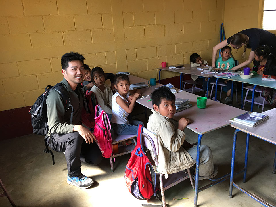 Man and children seated at a school desk in front of a chalkboard, with a classroom setting behind them.