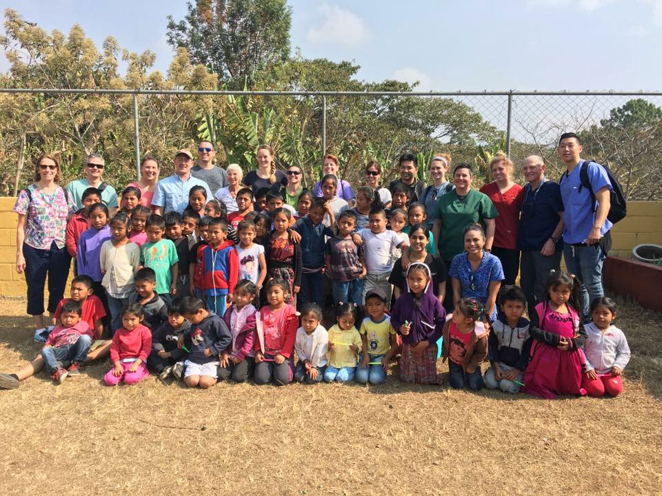 The image shows a group of people, including both adults and children, posing for a photo in an outdoor setting. They are standing on what appears to be a dirt ground with a fence in the background, possibly indicating a boundary or a safe area. The environment suggests a rural or semi-rural location.