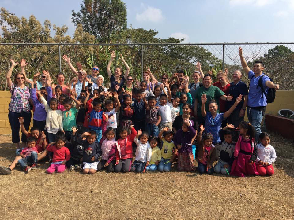 The image is a group photo of people, likely taken at an outdoor event or gathering in a dirt area with a fence in the background.