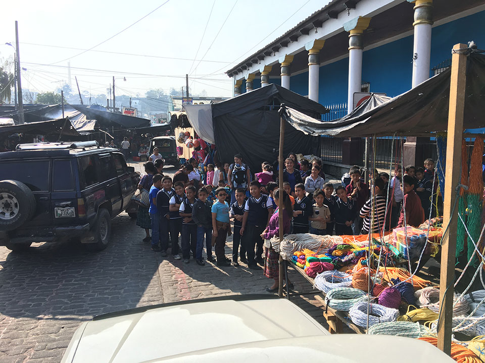 A group of people gathered at an outdoor market, with stalls displaying various items for sale.