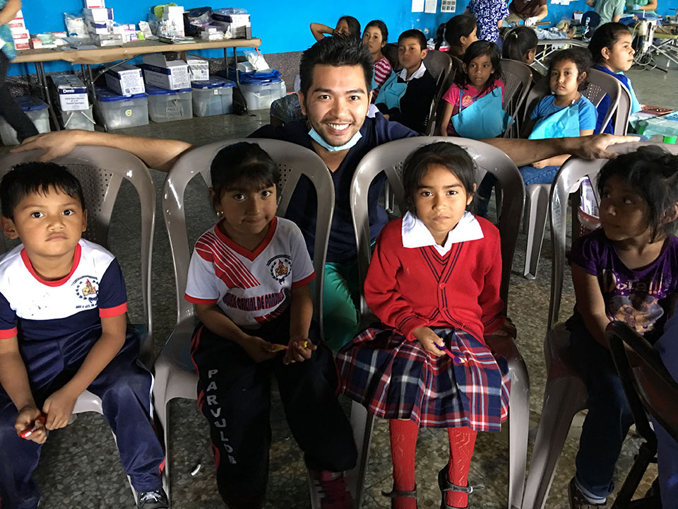 The image shows a man seated at the front of a room with children sitting in rows facing him. The man appears to be engaged in an activity or discussion with the children.