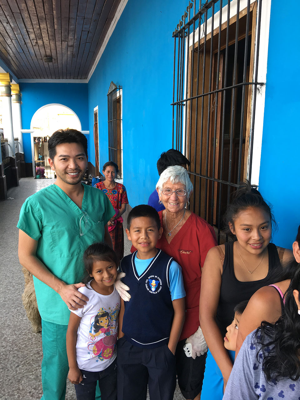 The image shows a group of people, including an individual wearing scrubs and a stethoscope, posing for a photo in front of a building with a blue door.