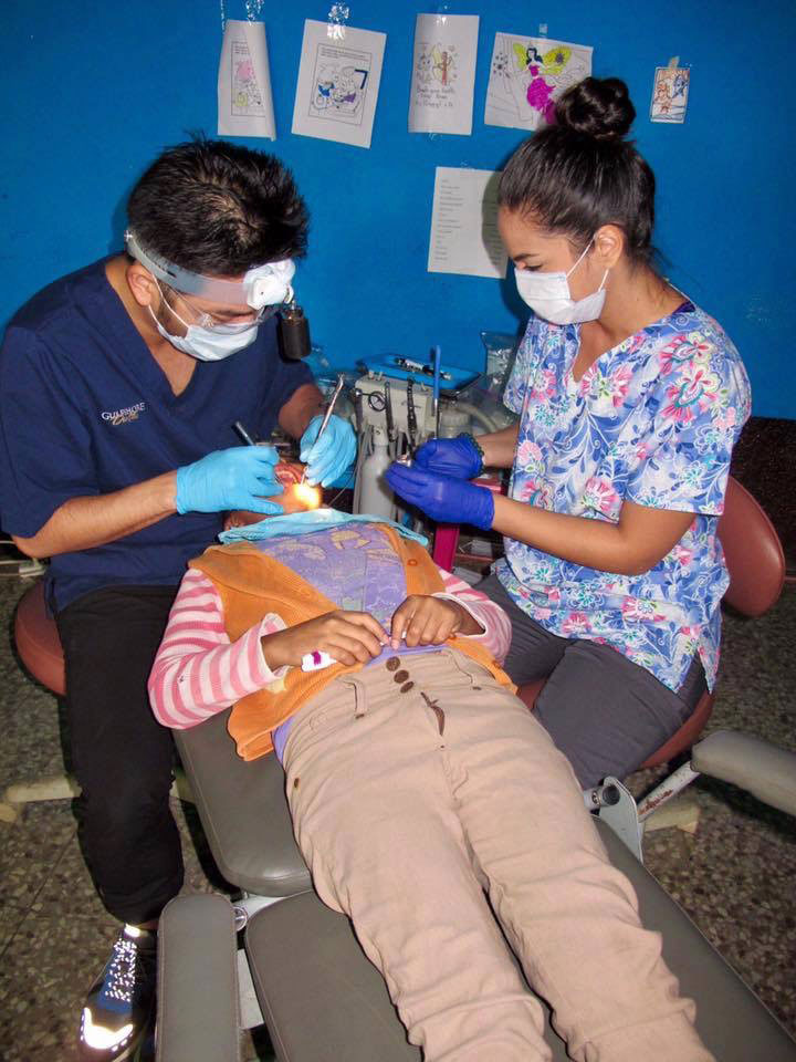 A dental office setting with a patient seated in a chair, receiving dental care from a dentist and dental hygienist.