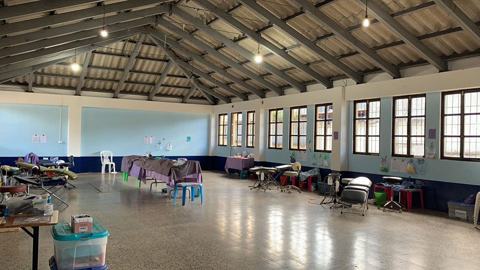 The image shows an interior space, likely a room in a facility, equipped with various pieces of furniture and equipment, including chairs, tables, mattresses, and what appears to be medical or therapy-related items. The floor is covered with white tiles, and there are large windows allowing natural light into the room.