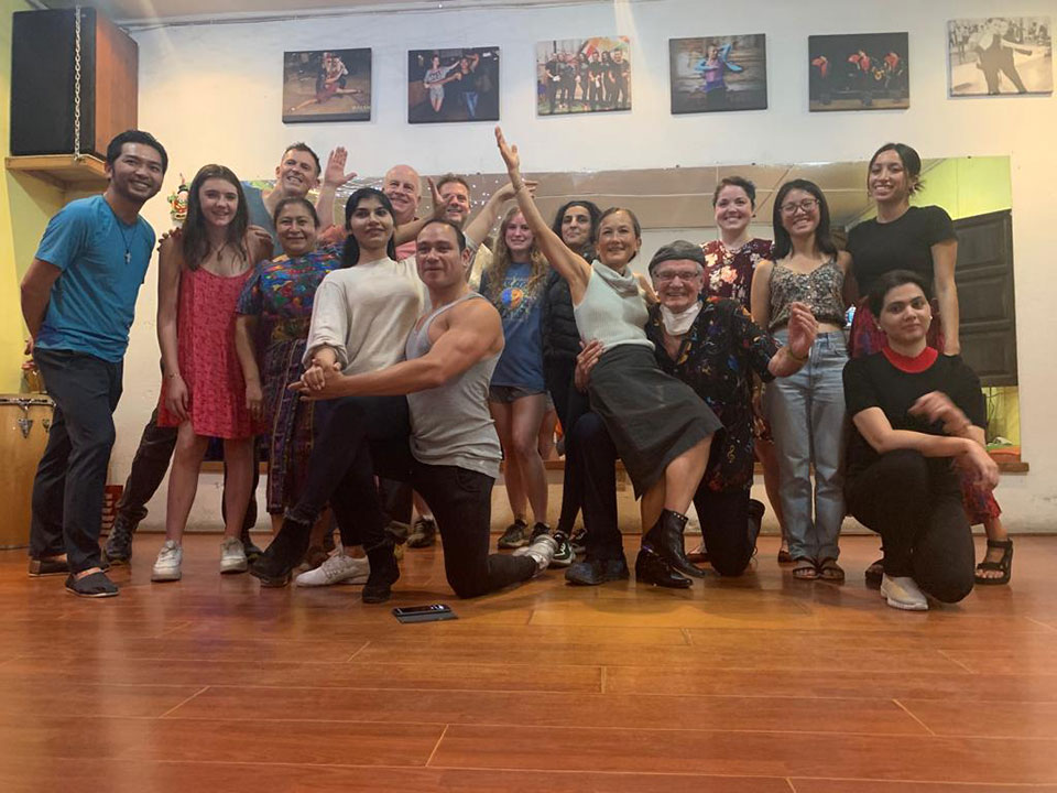 In the image, a group of people is posing for a photo in front of a stage with posters. The group consists of individuals of various ages and genders, some are smiling while others are making gestures. They appear to be gathered for an event or performance.