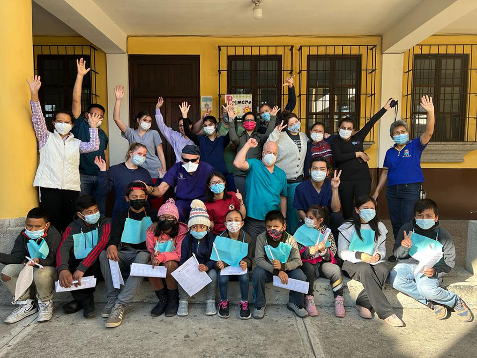 A group of individuals, both adults and children, wearing face masks and standing in front of a building with one hand raised, posing for a photograph.