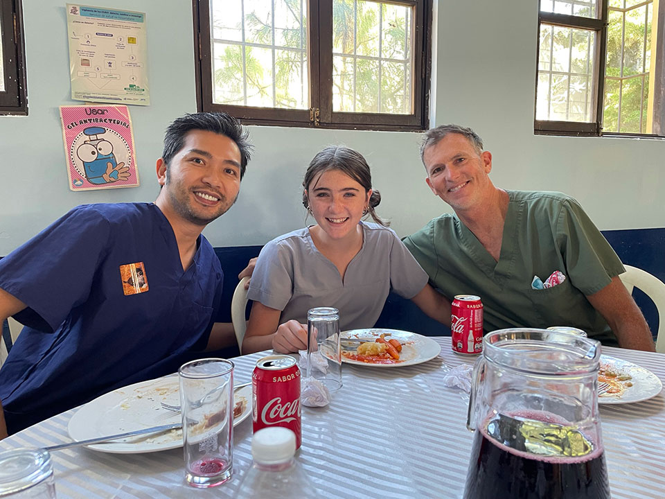 A group of three people, including a man in scrubs, sitting at a dining table with plates of food and drinks.