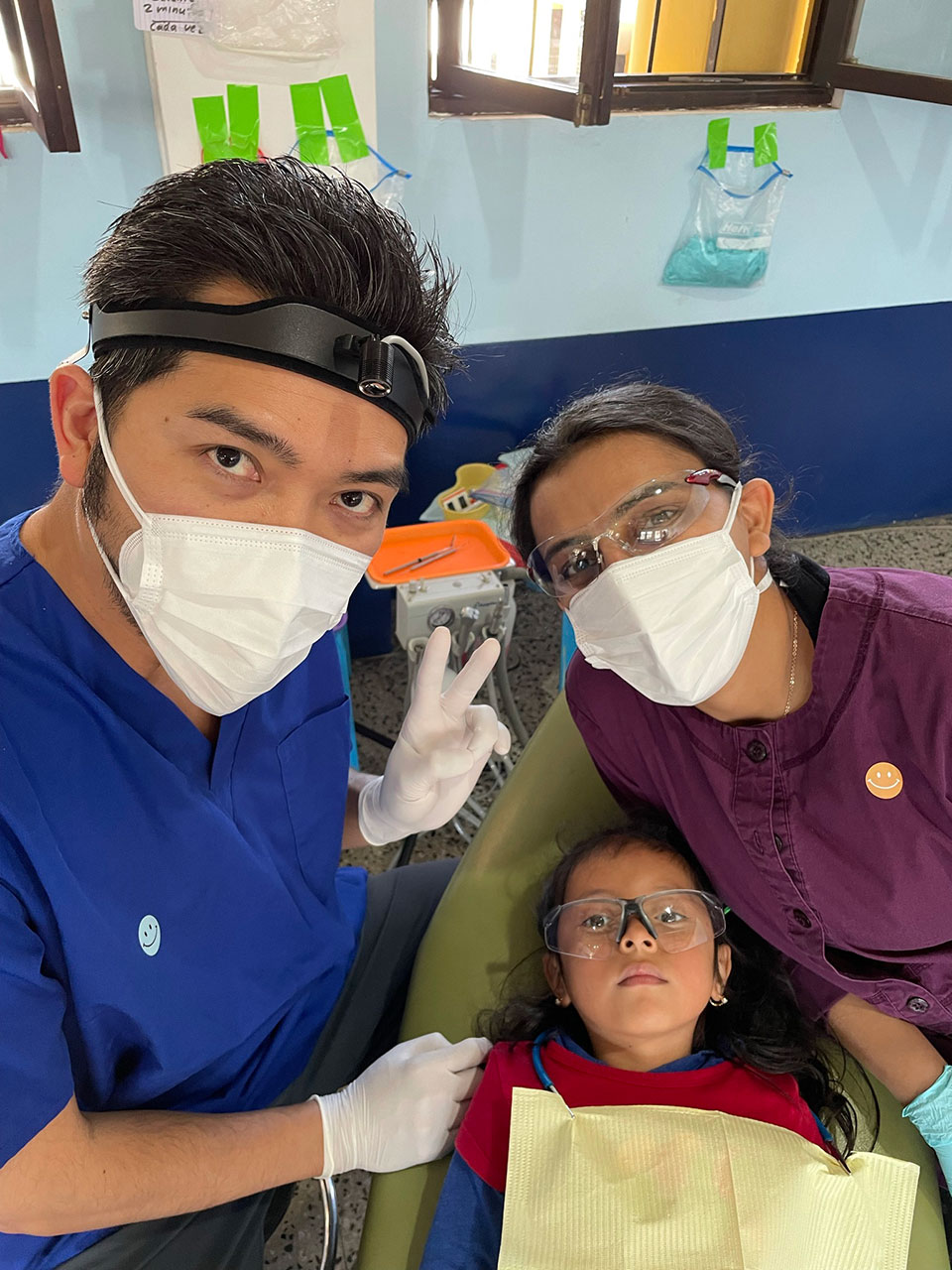 In the image, a medical professional in a mask is assisting a child wearing glasses and a face mask. They are both in a dental or medical setting, with a woman observing from behind. The adult appears to be taking care of the child's dental needs, as suggested by the presence of a toothbrush.