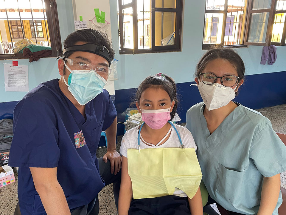 The image features three individuals in a room, with one person wearing a surgical mask and another person seated behind them. There's a child standing between the two adults. They are all dressed in casual clothing, suggesting an informal setting.