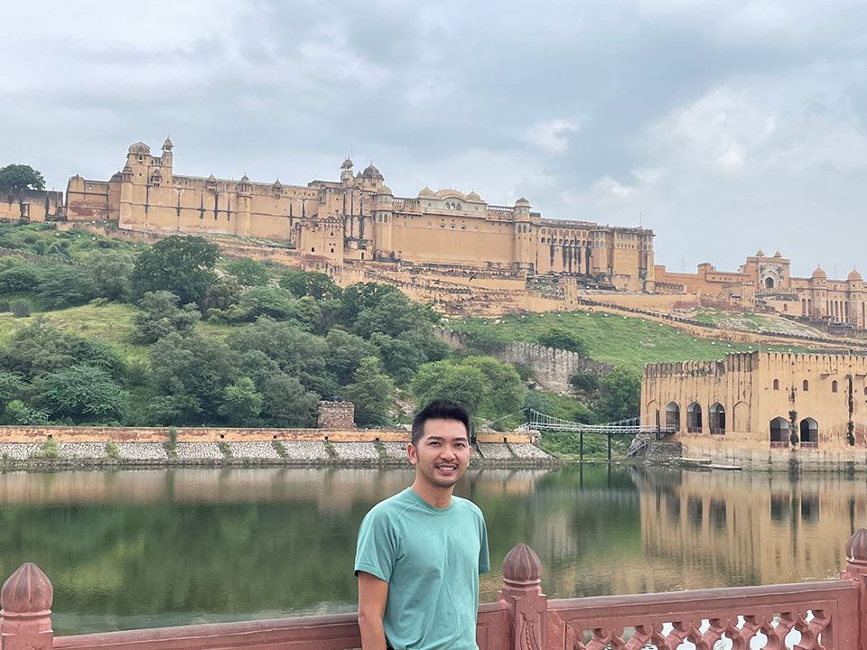 A man posing in front of a historic fort and palace complex with a scenic backdrop.