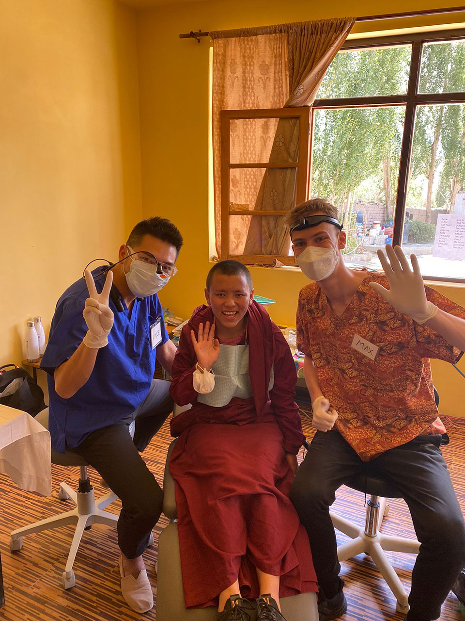 Three individuals, two men and a woman, wearing face masks are posing for the camera in front of a person sitting in a dental chair.