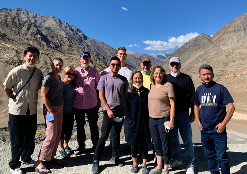 The image shows a group of people standing together outdoors, likely on a trip or tour, with a scenic mountainous landscape in the background.