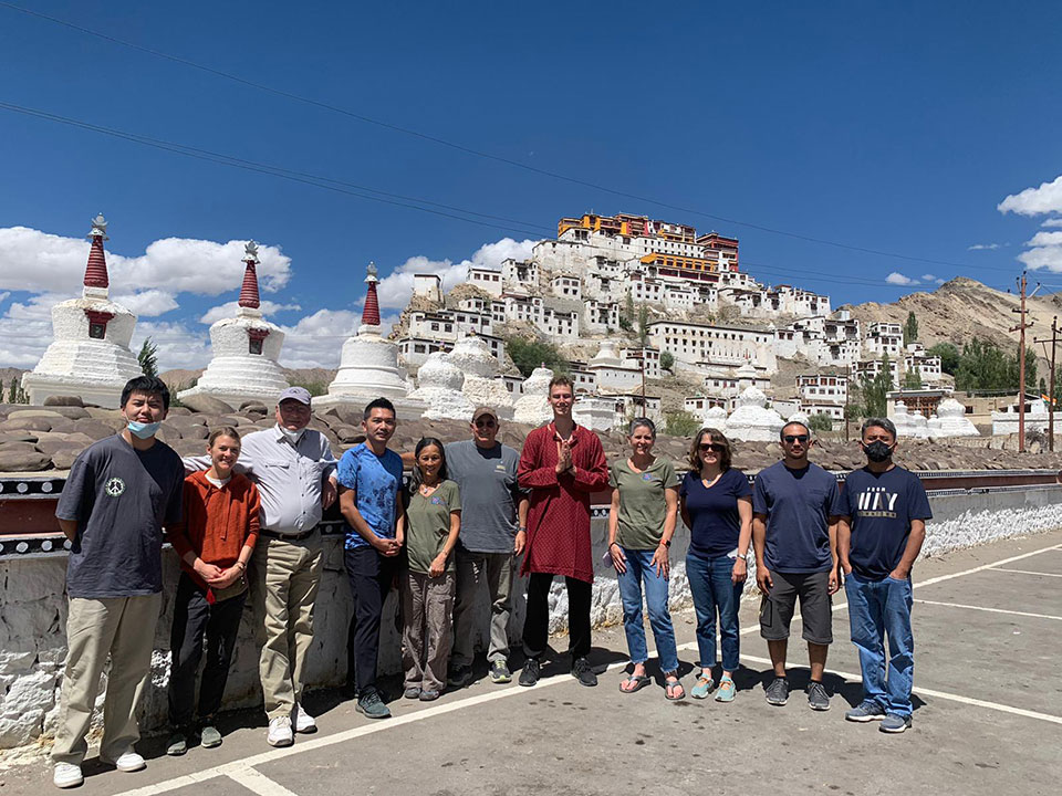 The image depicts a group of people standing in front of a large, ornate building with multiple white domes and red roofs, which is identified as the Potala Palace in Lhasa, Tibet.