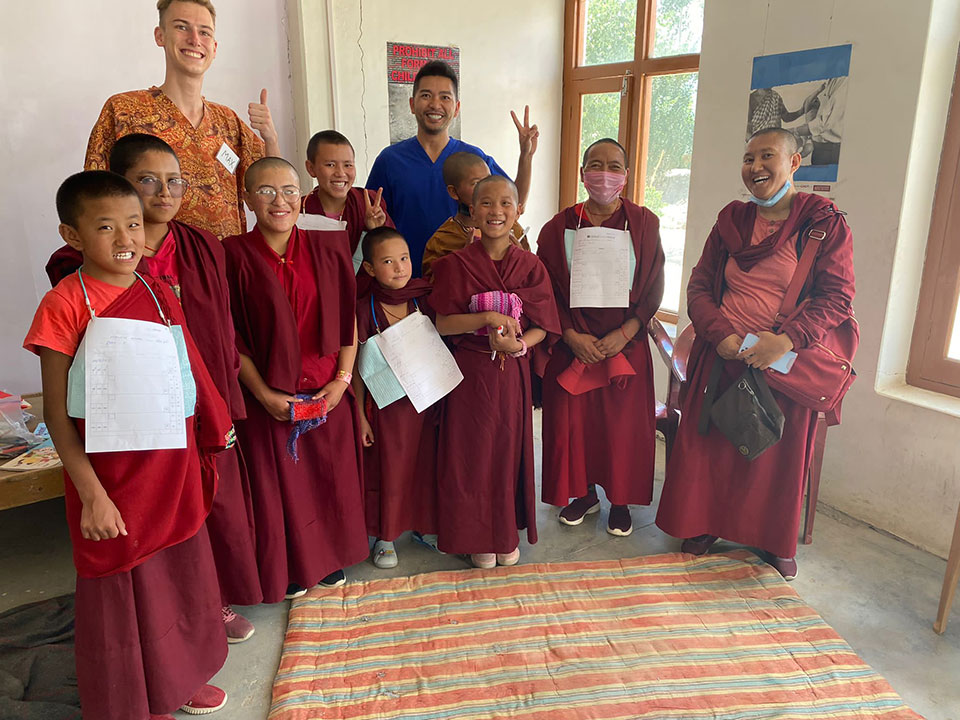 The image shows a group of people, likely Buddhist nuns, posing for a photo together. They are dressed in traditional monastic robes and appear to be in an indoor setting with a sign on the wall behind them.