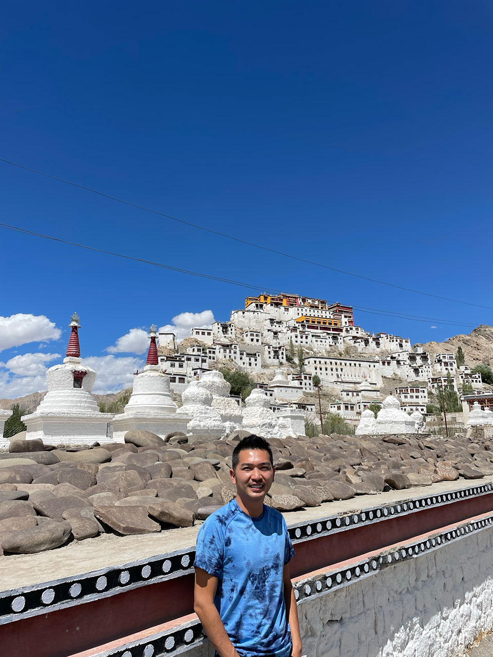 A person standing in front of a large, colorful Buddhist monastery with multiple buildings and a clear blue sky.