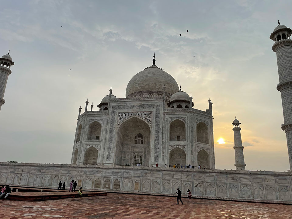 The image shows the Taj Mahal, a large white marble mausoleum with multiple domes and minarets, set against a hazy sky during what appears to be either sunrise or sunset.