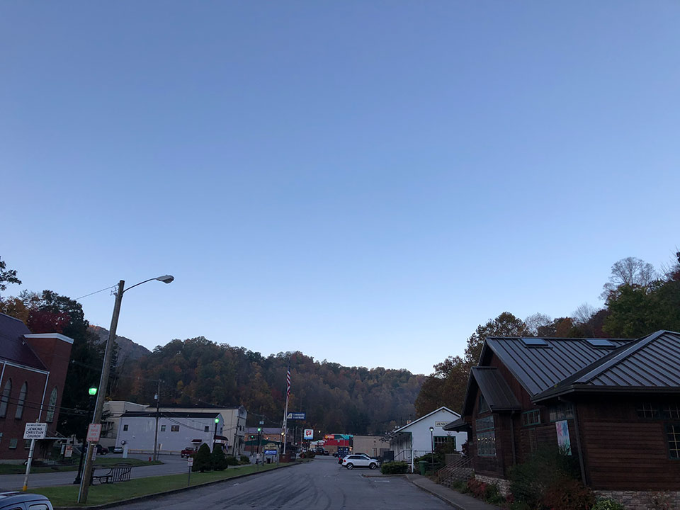 The image shows a serene street scene with a clear blue sky, trees in the background, and a road leading into a small town or village.