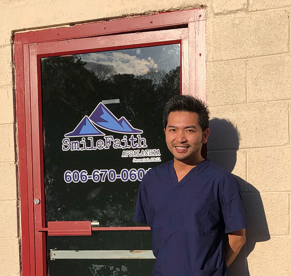 A smiling man in a white lab coat stands in front of a door with a sign that reads 'SMILEFAITH' and includes a phone number.