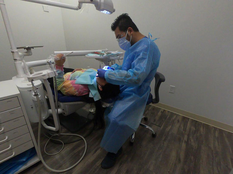 The image shows a dental professional in protective gear, sitting at a dental chair with equipment around them.