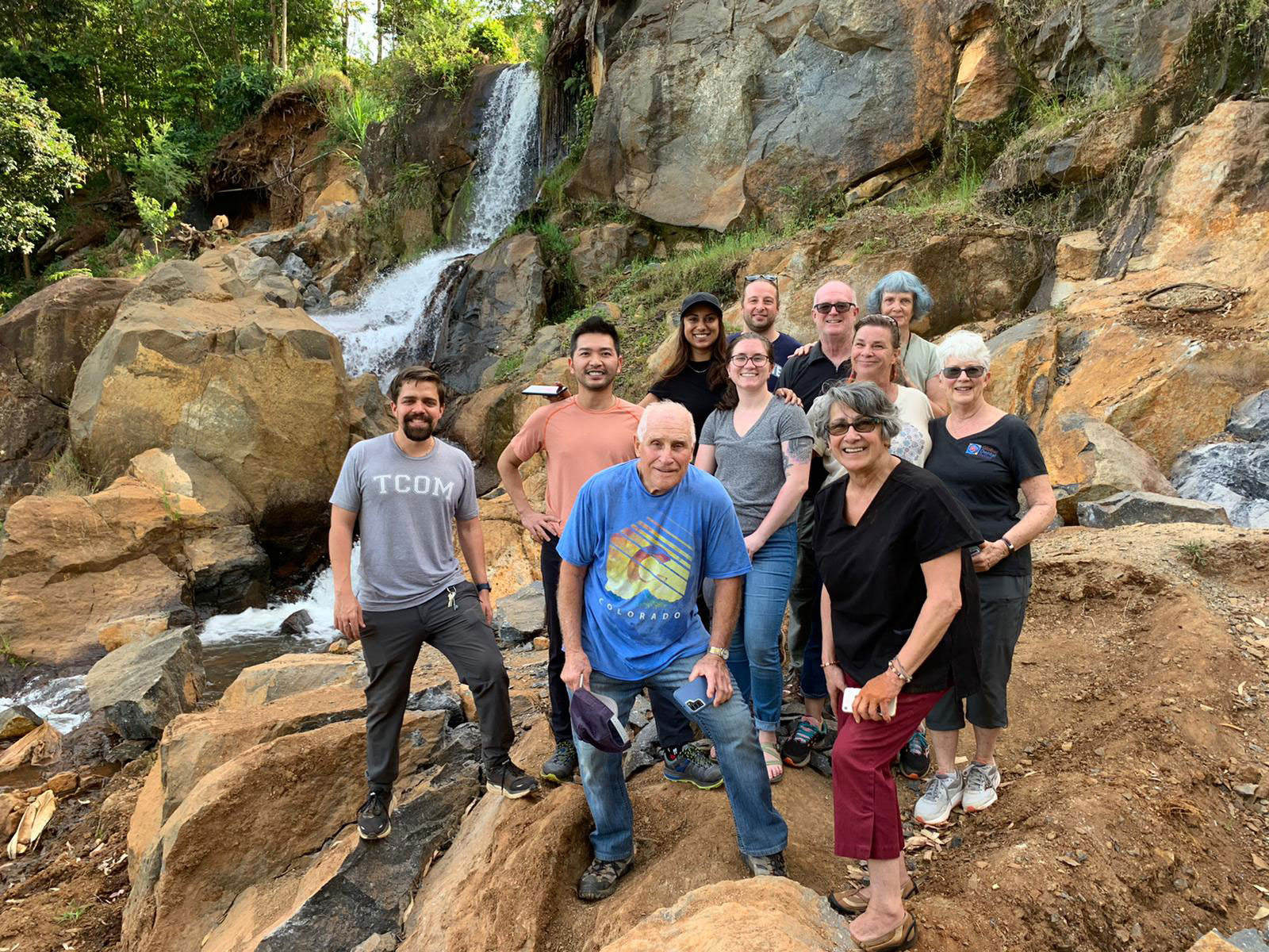 A group of people posing for a photo in front of a waterfall, with some standing on rocks and others on the ground.
