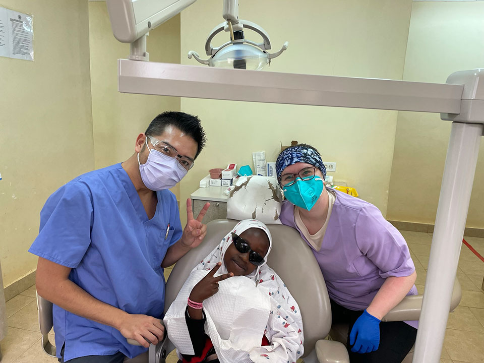 The image shows three individuals, likely healthcare professionals, standing in front of a dental chair with a patient. They are wearing face masks and one person is holding up a peace sign.