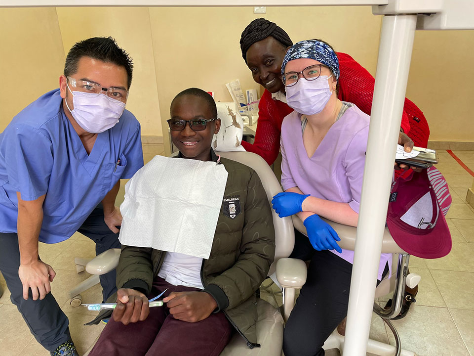 A group of healthcare professionals, including a man in a chair with a surgical mask on his face, posing for a photo with a smiling demeanor.
