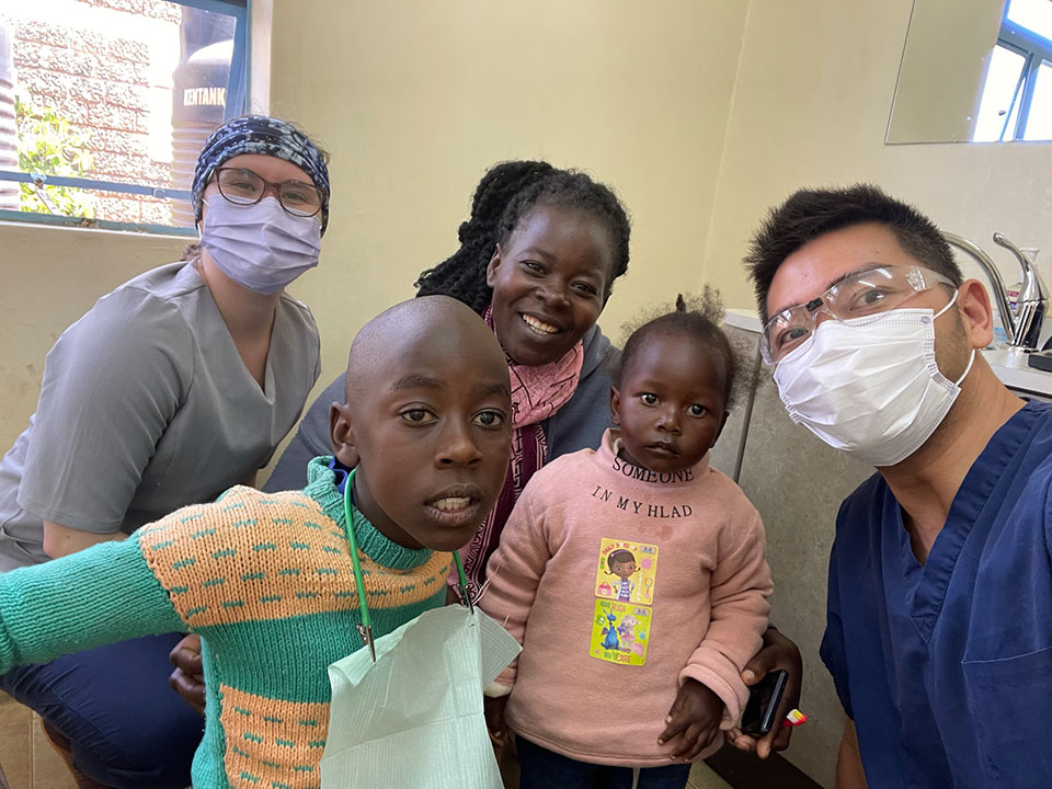 The image shows a group of people, including a healthcare worker wearing a face mask and surgical cap, posing in front of a dental chair with a young child. They are standing indoors, and there is a person taking the photograph.