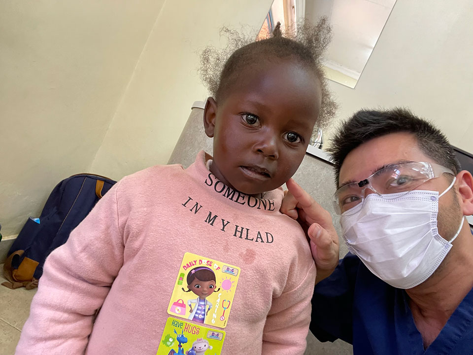 In the image, there is a young child with a sad expression looking at the camera, standing next to an adult wearing a white face mask and holding a stethoscope. The child has a small sticker on their shirt that reads 'I'm not my head.